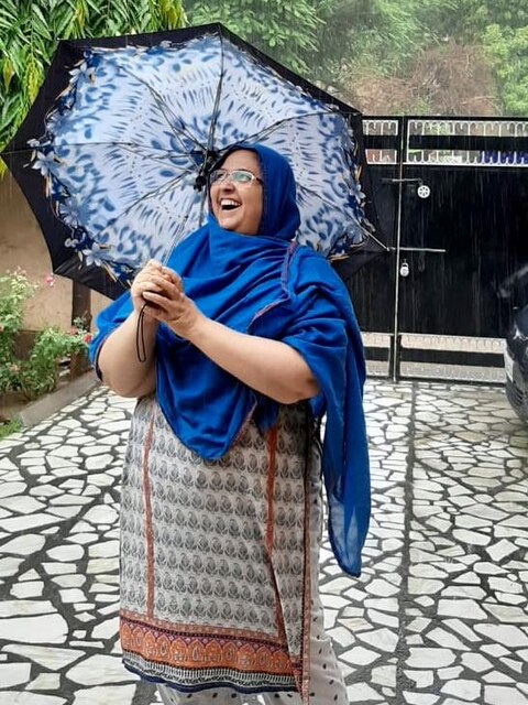 Badi Mami laughing in the rain under a blue umbrella, a Lucknow clock tower behind her